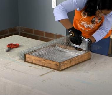 woman uses a stapler to attach hardware cloth to a wooden frame.